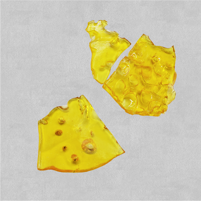 Three broken pieces of translucent amber shatter concentrate with a glassy, bubbled texture, on a light gray background.