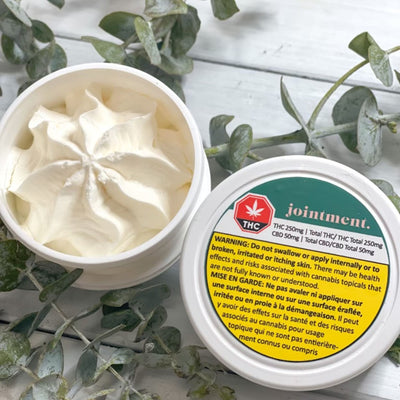 Open tub of whipped cream-colored Jointment massage butter beside its lid, styled with eucalyptus leaves.