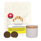 Cream-colored mylar package labeled Carmel Traditional Hash with a THC warning icon, next to loose hash pieces and a small ceramic storage jar with a wooden lid.