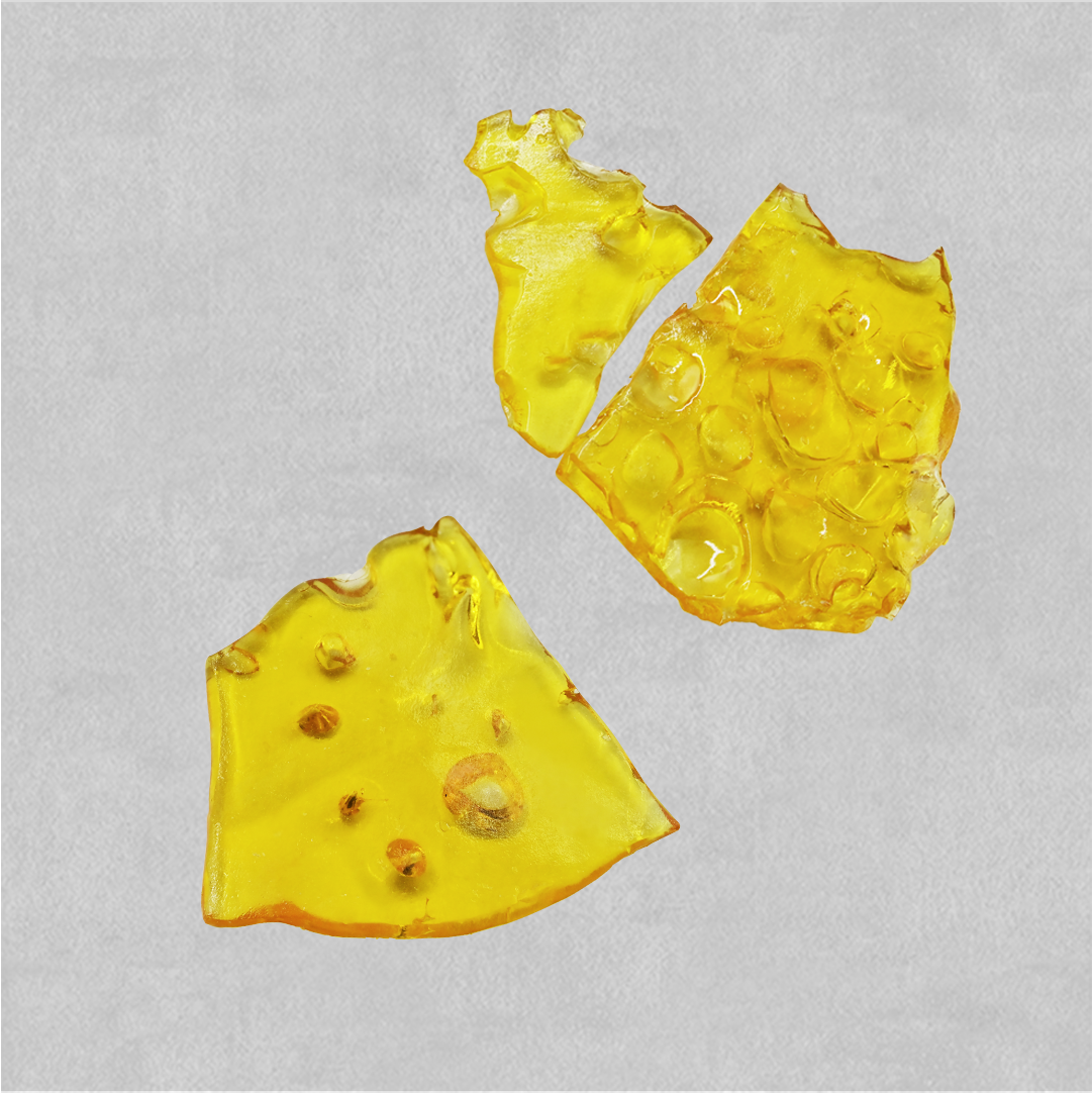 Three broken pieces of translucent amber shatter concentrate with a glassy, bubbled texture, on a light gray background.