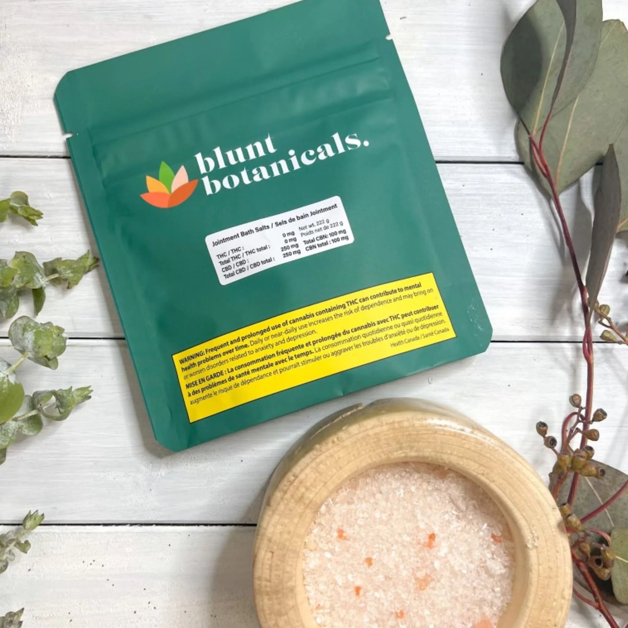 Teal packaging bag for Jointment Bath Salts by Blunt Botanicals next to a wooden bowl of pink bath salts, styled with eucalyptus leaves.